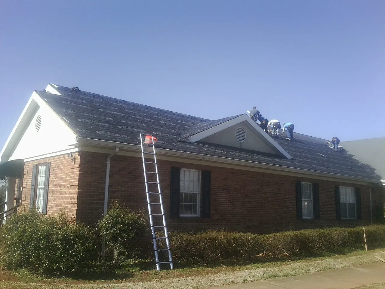 Crew tearing shingles off a brick ranch roof with a ladder set — Metal Roof Repair work by our crew in Straughn