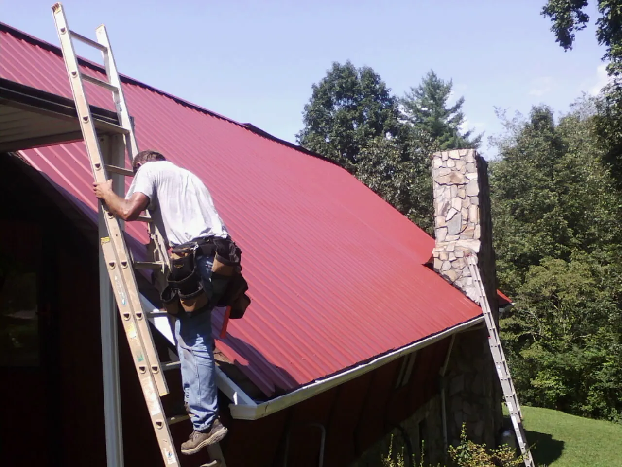 Roofer on a ladder beside a red metal roof and stone chimney — Soffit & Fascia Repair by Roofing Brothers in Straughn